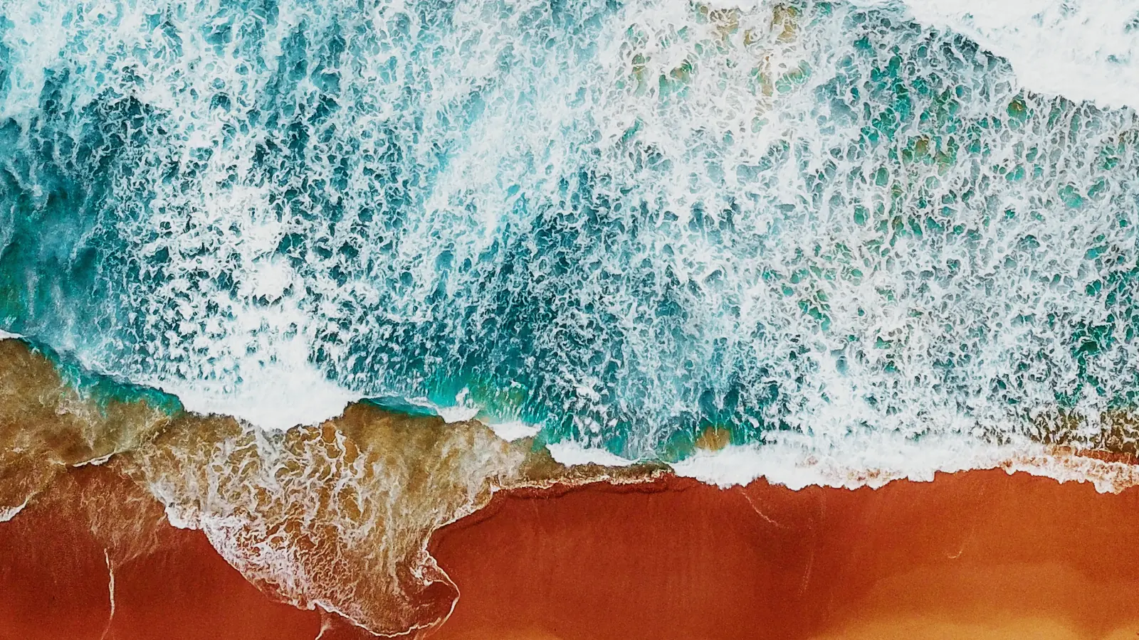 Surf breaking on a red-sand beach, seen from above.