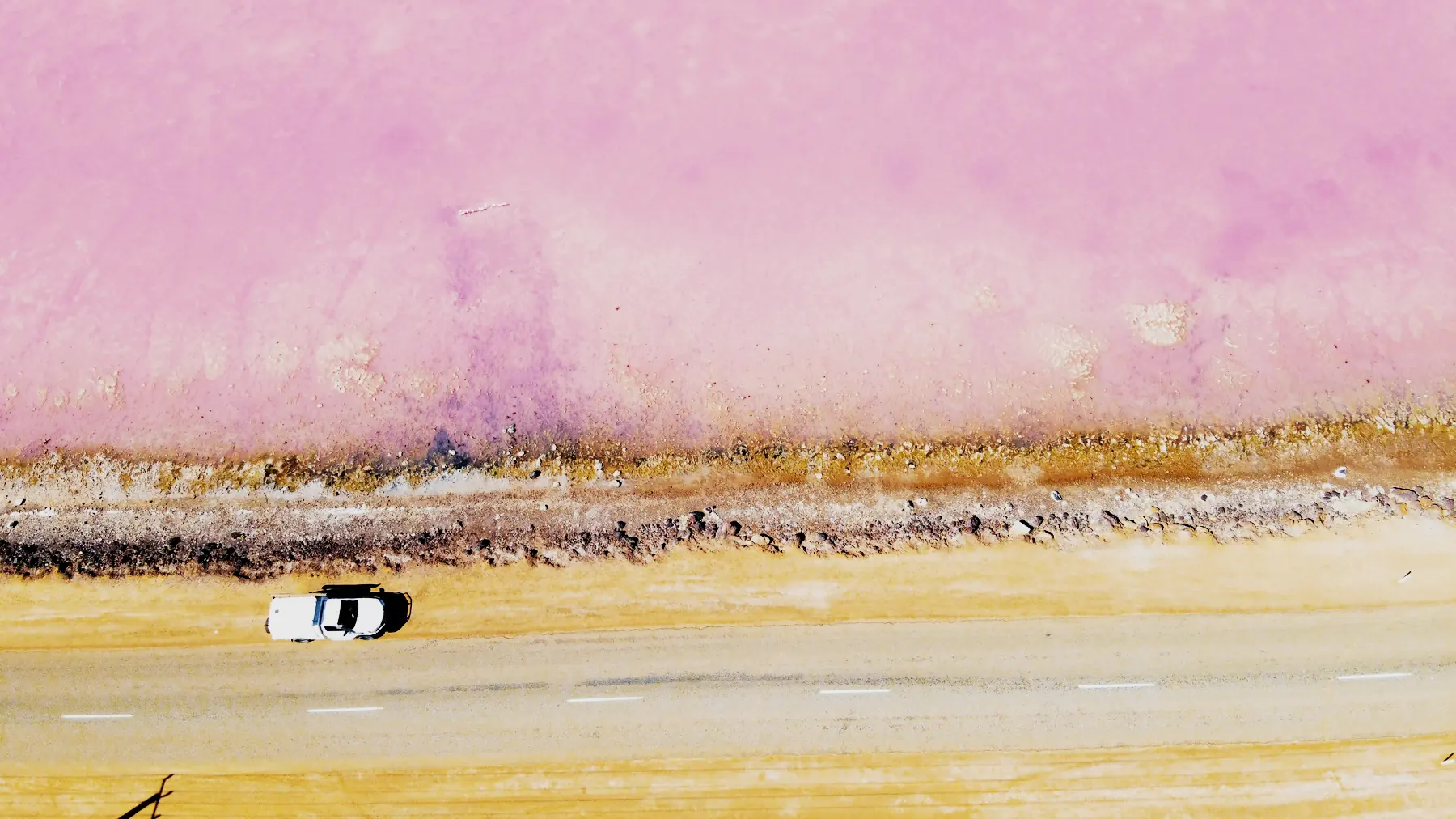A pink salt lake beside a desert highway in Western Australia, seen from above.