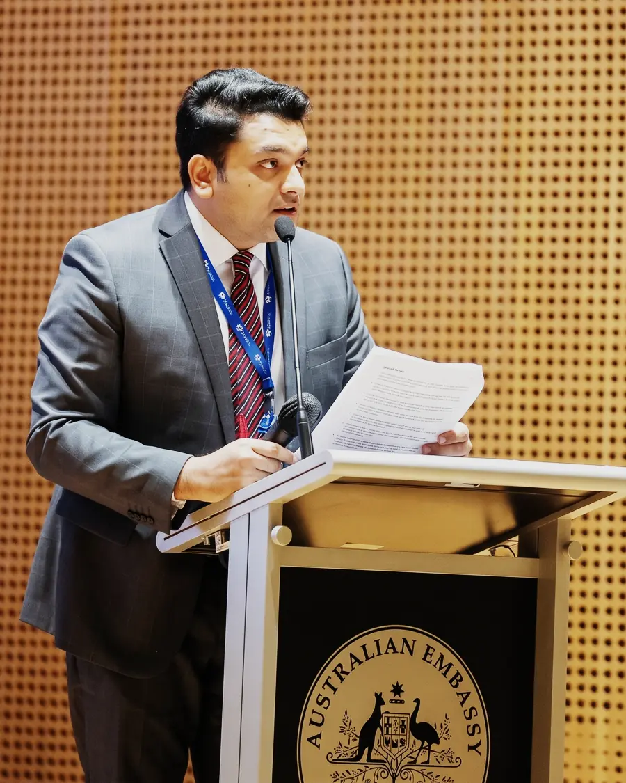 Aqeeb Akram speaking from a lectern bearing the Australian Embassy crest.